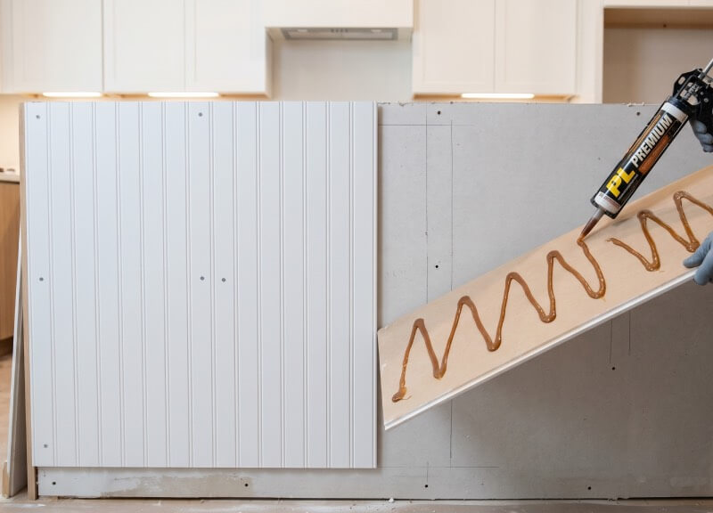 A DIYer applying a zigzag pattern of PL Premium polyurethane construction adhesive to the back of a beadboard panel for a kitchen island.