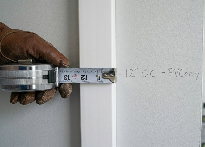 Close-up showing a hand measuring a white PVC batten strip on a painted wall for a DIY bathroom board-and-batten project, with a pencil annotation showing precise spacing.