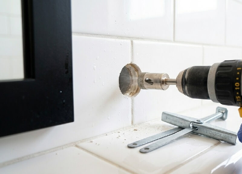 Close-up of a drill with a diamond core bit creating a clean hole through white porcelain tile for installing a heavy-duty mirror anchor during a DIY bathroom remodel.