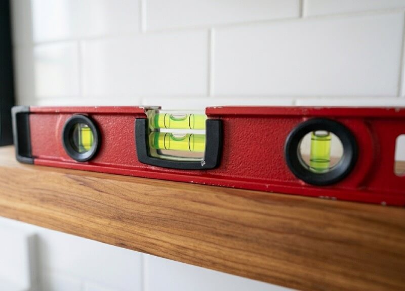 Torpedo level resting flat on a custom cedar floating shelf installed directly over a white subway tile bathroom backsplash.