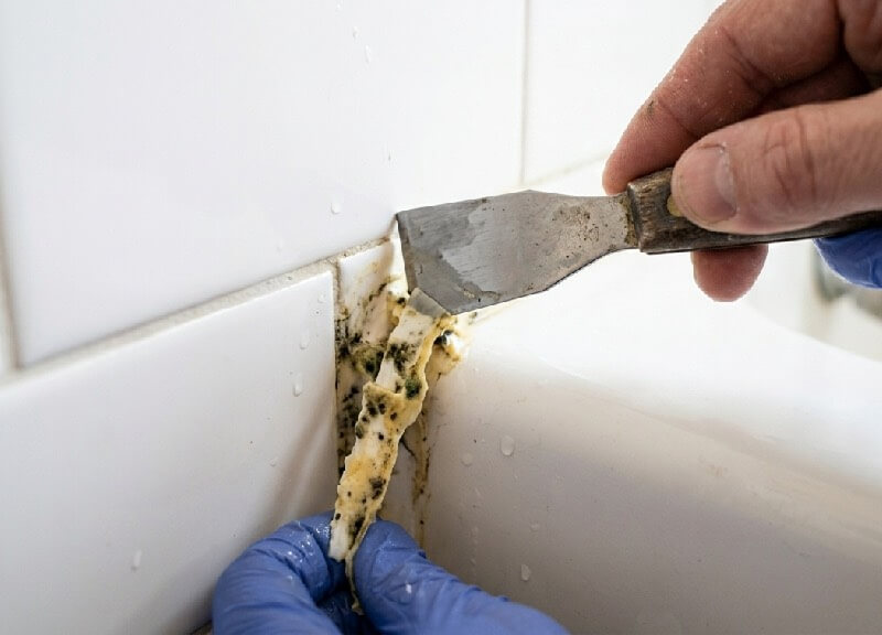 Extreme close-up of a metal scraping tool removing old, yellowed, mildew-infested silicone caulk from a white tile shower corner during a DIY bathroom remodel.