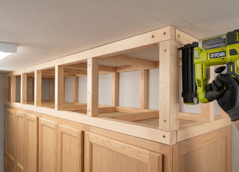 Close-up of a gloved hand using a Ryobi brad nailer to assemble a raw wood soffit skeleton above light oak kitchen cabinets.