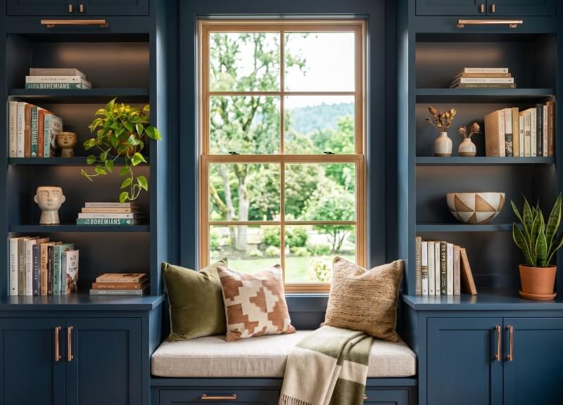 Dark navy blue built-in bookshelves with copper hardware and a cozy window seat framing a bright window.