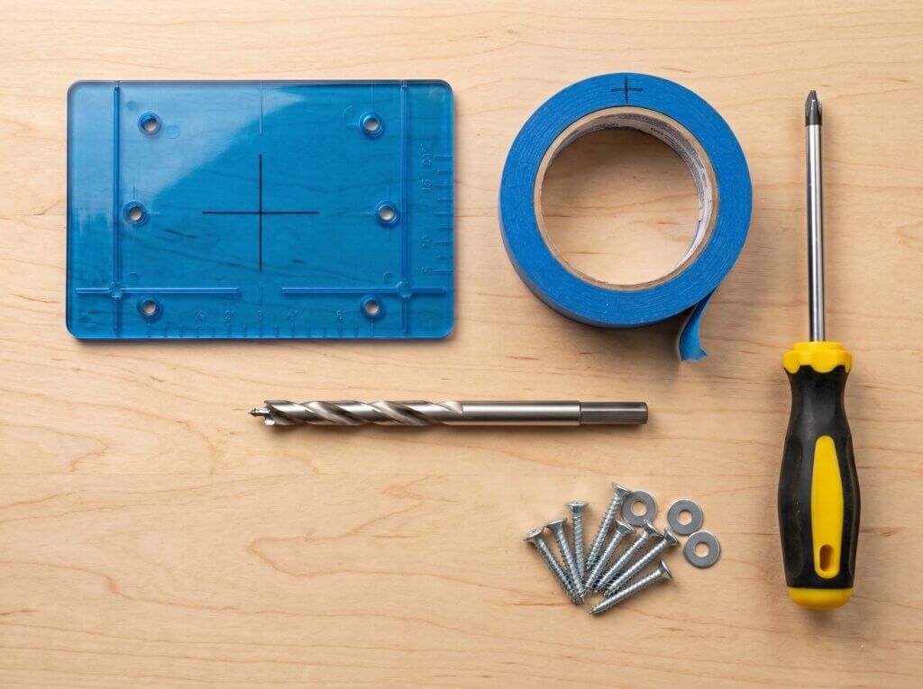 A flat lay of cabinet hardware installation tools on a wooden workbench, showing the blue placement template, the blue masking tape (with a drawn crosshair), the manual screwdriver, the hardware pile, and a correctly detailed brad-point drill bit with its central spike.